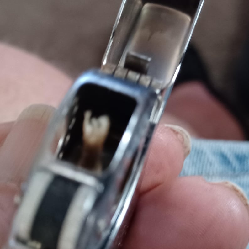 May include: A silver-toned metal lighter with the lid flipped open, revealing a hollow interior chamber. The casing features a smooth, reflective finish with a dark, textured vertical band running along the side of the main body structure.