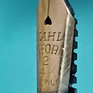 May include: Close-up of a gold-colored fountain pen nib. The nib has the text "AHL FOR 2 4K EINUSA" engraved on it. The nib has a heart-shaped breather hole and is set against a blue background.