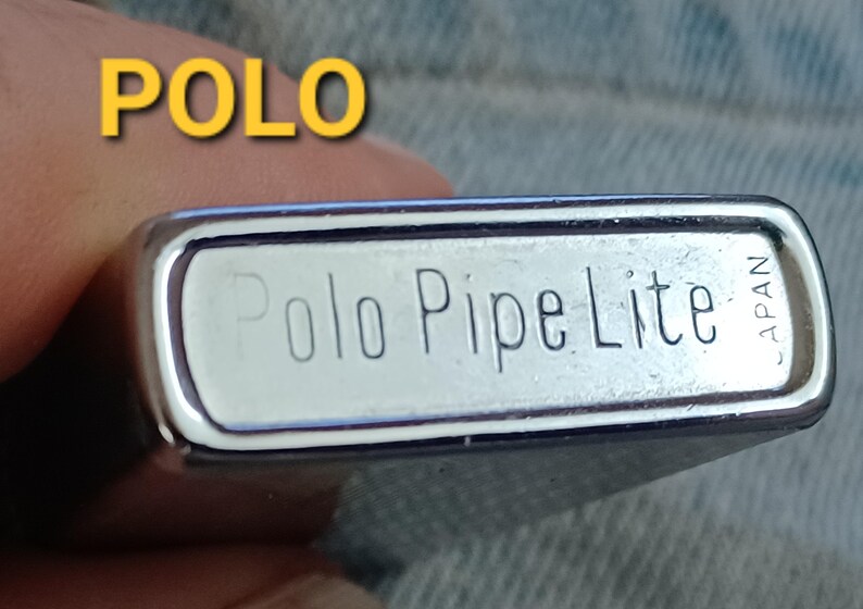 May include: A rectangular silver-toned metal lighter with rounded edges. The bottom surface is engraved with the text 'Polo Pipe Lite' and 'JAPAN'. The word 'POLO' is also displayed in yellow text in the upper left corner of the image.