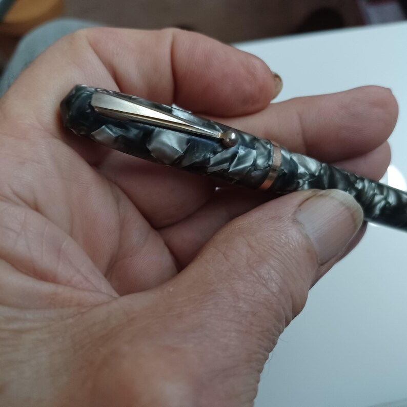 May include: A luxury fountain pen with a marbled gray and black body, gold-colored clip, and trim. The pen is held in a hand, showcasing its elegant design and craftsmanship. The pen's cap features a gold-colored clip.