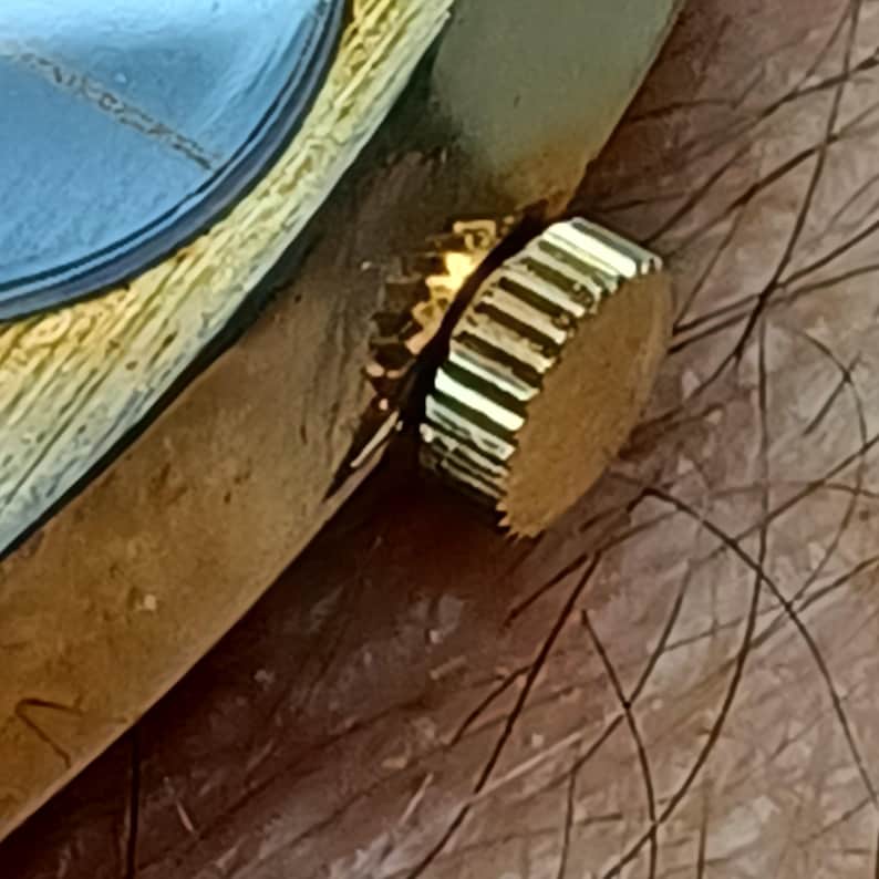 Puede incluir: Primer plano de una corona de reloj dorada. La corona es cil&iacute;ndrica con un borde estriado para agarre y est&aacute; colocada sobre una superficie marr&oacute;n. La esfera del reloj es visible en el fondo.