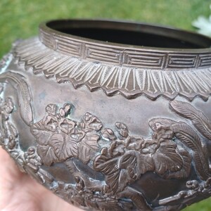 May include: A round, bronze planter with intricate floral and leaf designs. The planter has a raised rim with a geometric pattern.