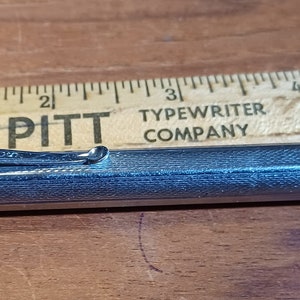 May include: A silver retractable pen with a red and white stripe on the end. The pen is lying on a wooden ruler with the text "FORT PITT TYPEWRITER COMPANY 644 LIBERTY AVE. PITTSBURGH 22, PA. ATLANTIC 1-8320".