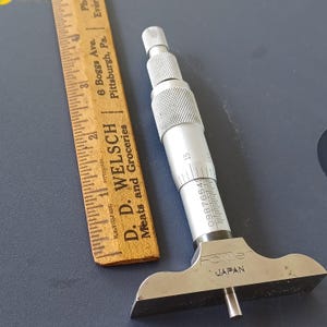 May include: A silver Fowler depth micrometer with a knurled grip and a measurement scale. A wooden ruler with markings and text is also present. The ruler is marked with the text "D. D. Welsch Meats and Groceries".