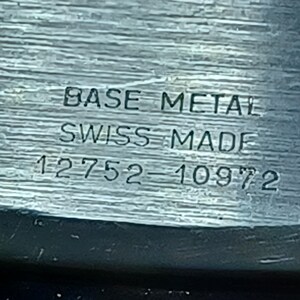 Puede incluir: Primer plano de un componente de reloj con el texto "BASE METAL", "SWISS MADE" y los n&uacute;meros "12752-10972" grabados en el metal. La superficie tiene un aspecto texturizado y cepillado.