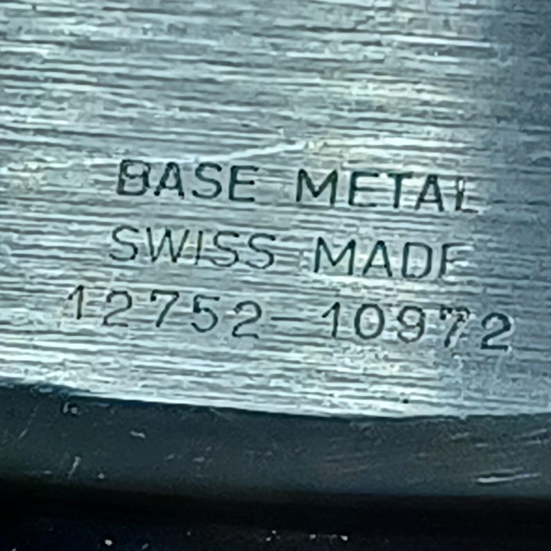Puede incluir: Primer plano de un componente de reloj con el texto "BASE METAL", "SWISS MADE" y los n&uacute;meros "12752-10972" grabados en el metal. La superficie tiene un aspecto texturizado y cepillado.