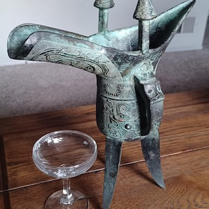 May include: A bronze Chinese ritual wine vessel with intricate carvings and three legs. The vessel is green and has a spout on one side. It is sitting on a wooden table next to a clear glass.