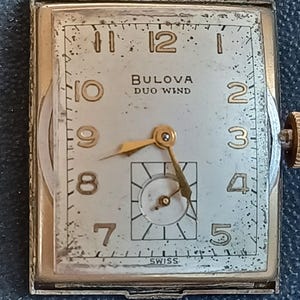 K&ouml;nnte beinhalten: Eine goldfarbene Armbanduhr mit quadratischem Zifferblatt. Das Zifferblatt ist wei&szlig; mit schwarzen Markierungen und goldfarbenen Zeigern. Die Uhr tr&auml;gt die Aufschrift "BULOVA DUO WIND" auf dem Zifferblatt. Die Uhr ist unten auf dem Zifferblatt mit "SWISS" gekennzeichnet.