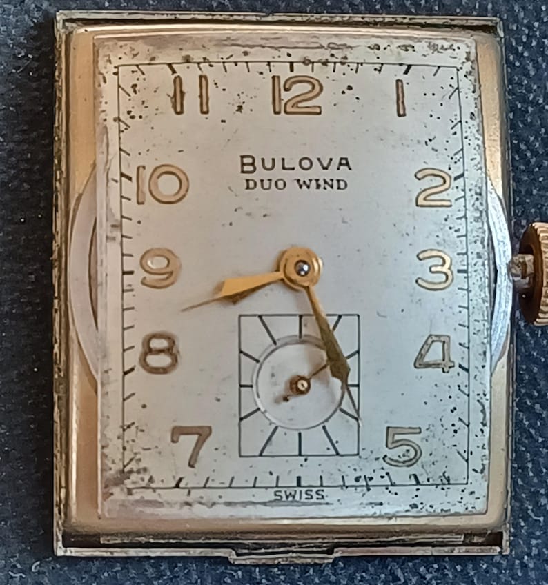 K&ouml;nnte beinhalten: Eine goldfarbene Armbanduhr mit quadratischem Zifferblatt. Das Zifferblatt ist wei&szlig; mit schwarzen Markierungen und goldfarbenen Zeigern. Die Uhr tr&auml;gt die Aufschrift "BULOVA DUO WIND" auf dem Zifferblatt. Die Uhr ist unten auf dem Zifferblatt mit "SWISS" gekennzeichnet.