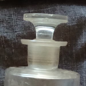 May include: A clear glass bottle with a stopper. The bottle has a round body and a cylindrical neck. The stopper is a simple, flat disc with a small lip.