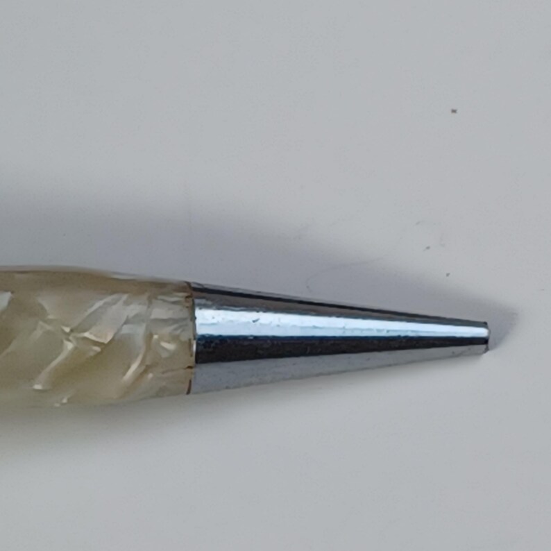 May include: Close-up of a silver mechanical pencil tip. The tip is a shiny, metallic cone shape. The pencil's body is a light, pearlescent colour. The image is set against a plain, off-white background.