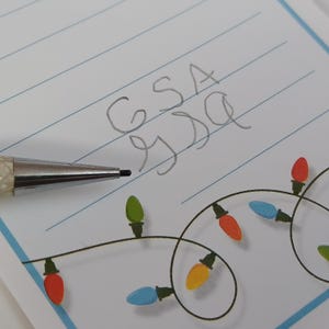 May include: A close-up of a lined notepad with the handwritten letters "GSA" and "SSD". A silver mechanical pencil is positioned near the text. The notepad features a decorative string of colourful Christmas lights along the bottom edge.