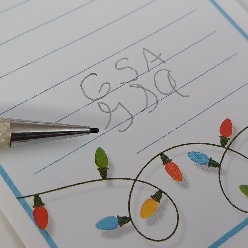 May include: A close-up of a lined notepad with the handwritten letters "GSA" and "SSD". A silver mechanical pencil is positioned near the text. The notepad features a decorative string of colourful Christmas lights along the bottom edge.