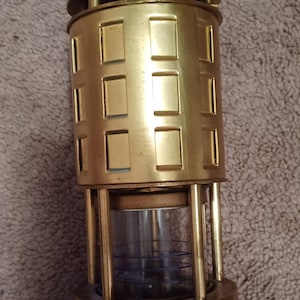 May include: A brass miner's lamp with a hook on top and a glass cylinder in the middle. The lamp has a decorative design with a series of rectangular cutouts.