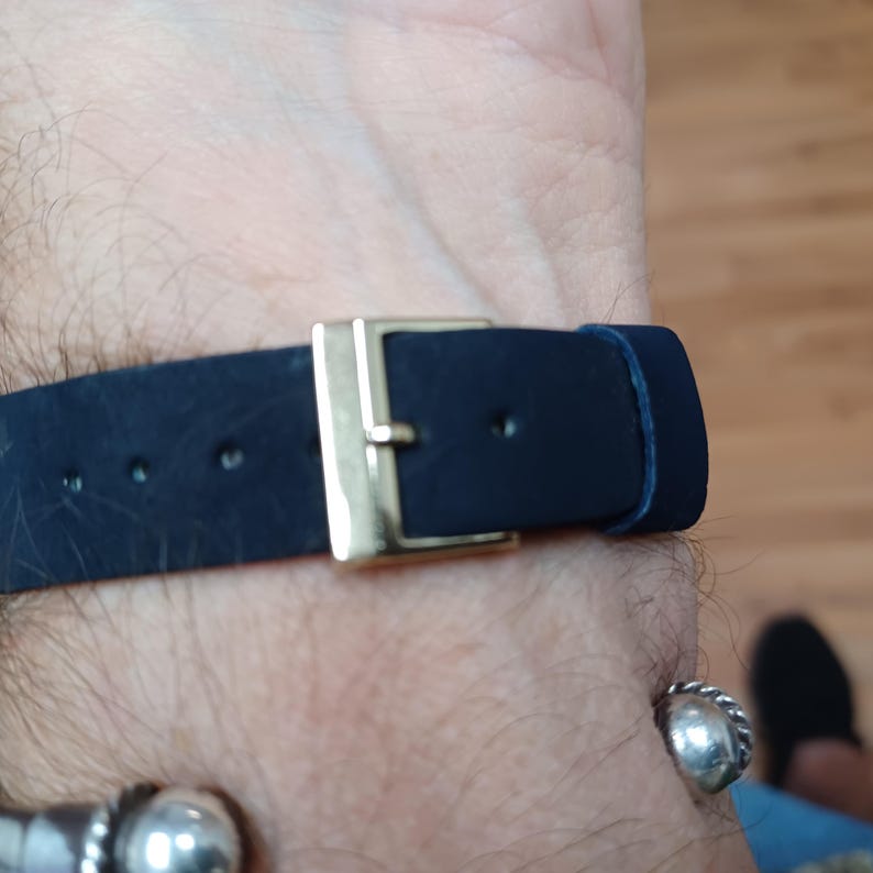 Puede incluir: Primer plano de una correa de reloj azul marino con una hebilla dorada. La correa es de cuero y tiene varios agujeros para ajustar. La hebilla es rectangular y brillante. La correa del reloj est&aacute; abrochada en una mu&ntilde;eca.