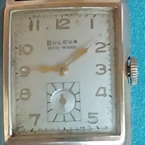 K&ouml;nnte beinhalten: Eine goldfarbene Bulova-Uhr mit wei&szlig;em Zifferblatt und schwarzem Lederarmband. Die Uhr hat ein rechteckiges Zifferblatt mit schwarzen r&ouml;mischen Ziffern als Stundenmarkierungen und einem goldenen Sekundenzeiger. Das Zifferblatt der Uhr tr&auml;gt au&szlig;erdem den Text "Bulova Self Wind".