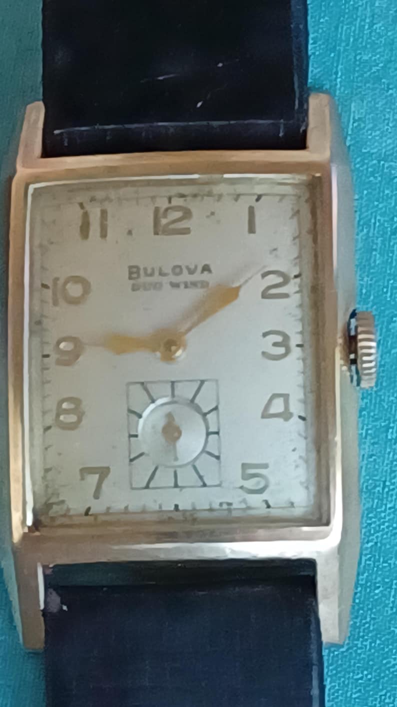 K&ouml;nnte beinhalten: Eine goldfarbene Bulova-Uhr mit wei&szlig;em Zifferblatt und schwarzem Lederarmband. Die Uhr hat ein rechteckiges Zifferblatt mit schwarzen r&ouml;mischen Ziffern als Stundenmarkierungen und einem goldenen Sekundenzeiger. Das Zifferblatt der Uhr tr&auml;gt au&szlig;erdem den Text "Bulova Self Wind".