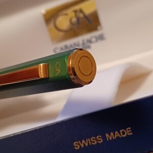 Puede incluir: Primer plano de una pluma verde con detalles dorados. La pluma tiene la palabra "Caran d'Ache" grabada en el lateral y las palabras "Swiss Made" en la caja.