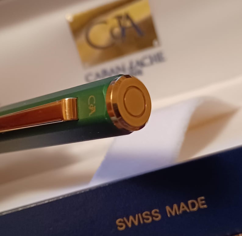 Puede incluir: Primer plano de una pluma verde con detalles dorados. La pluma tiene la palabra "Caran d'Ache" grabada en el lateral y las palabras "Swiss Made" en la caja.