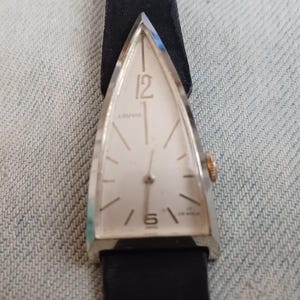 May include: A triangular-shaped wristwatch with a silver-toned case and a white dial. The face features the text LOUVIC, the number 12 at the top, the number 6 at the bottom, and the text 17 JEWELS. It has a black strap and a gold-toned crown.