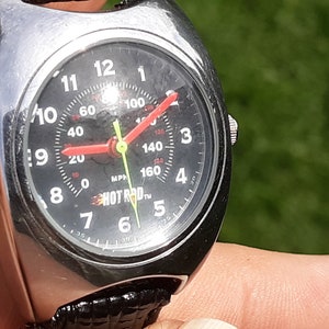 May include: A silver-toned wristwatch with a black face featuring white numerals and a red speedometer needle. The watch face includes the text "HOT ROD" and a speedometer that measures up to 160 MPH. The watch has a black leather band.