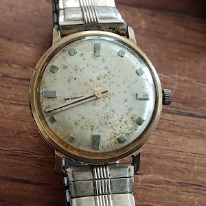 May include: Vintage gold-toned wristwatch with a silver-colored band. The watch face is off-white with rectangular hour markers and black hands. The watch shows signs of age and wear, with a tarnished appearance.
