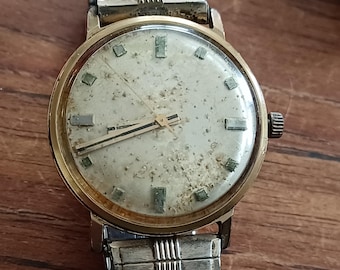 Op de afbeelding: Vintage goudkleurig polshorloge met een zilverkleurige band. De wijzerplaat is gebroken wit met rechthoekige uurmarkeringen en zwarte wijzers. Het horloge vertoont tekenen van ouderdom en slijtage.