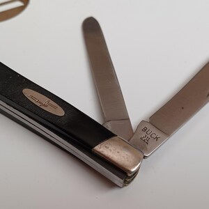 May include: A folding pocket knife with a black textured handle and silver metal bolsters. The knife features two extended stainless steel blades, one of which is stamped with the text BUCK and a symbol. The handle displays an oval metal inlay.