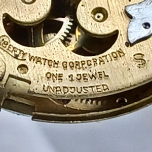 May include: Close-up of a vintage watch movement with visible gears and the text "LIBERTY WATCH CORPORATION." The watch mechanism is gold-toned with the text "ONE 1 JEWEL UNADJUSTED."