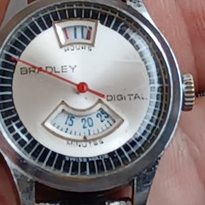 May include: A silver-tone Bradley Digital wristwatch with a white circular dial featuring a black grid border. The watch displays hours in a top window and minutes in a bottom window with blue numerals. A thin red hand extends across the center.