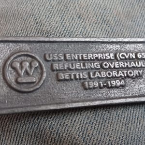 May include: A rectangular, silver-colored metal plaque with the text "USS ENTERPRISE (CVN 65) REFUELING OVERHAUL BETTIS LABORATORY 1991-1994". The plaque also features a circular emblem with a "W" design.