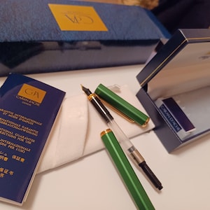 Puede incluir: Un bol&iacute;grafo fuente Caran d'Ache verde y dorado con un cartucho de tinta negro. El bol&iacute;grafo est&aacute; en su caja, que es azul con ribetes dorados. La caja tambi&eacute;n contiene un peque&ntilde;o folleto con instrucciones y una garant&iacute;a.
