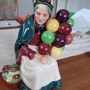 May include: A ceramic figurine of an elderly woman wearing a green shawl and a red dress, holding a bunch of colorful balloons.