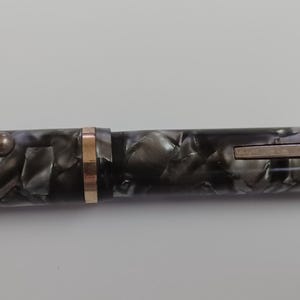 May include: A vintage fountain pen with a marbled black and gray barrel. The pen features gold-colored trim, including a clip and a band near the nib. The pen is a classic writing instrument.