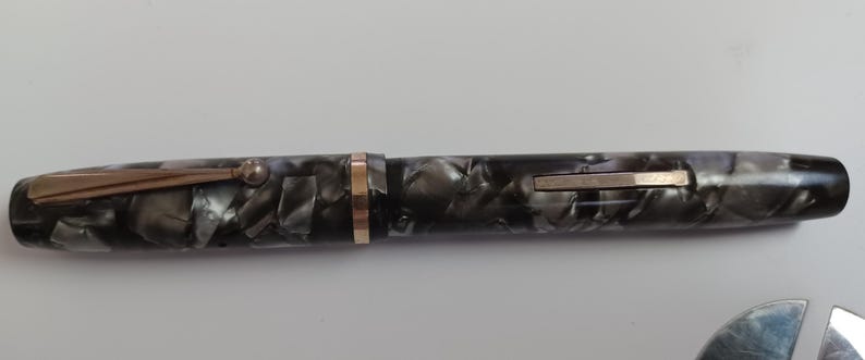 May include: A vintage fountain pen with a marbled black and gray barrel. The pen features gold-colored trim, including a clip and a band near the nib. The pen is a classic writing instrument.