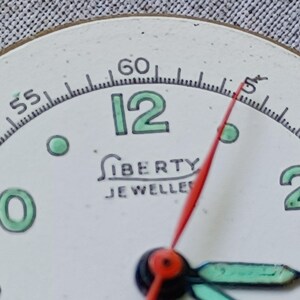 May include: Close-up of a vintage watch face with a white background. The number 12 is in green, and the hands are red and green. The text "LIBERTY JEWELLERS" is printed below the center.