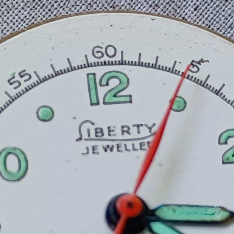May include: Close-up of a vintage watch face with a white background. The number 12 is in green, and the hands are red and green. The text "LIBERTY JEWELLERS" is printed below the center.
