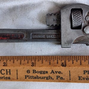 May include: An antique adjustable pipe wrench with a red stripe on the handle, next to a wooden ruler. The wrench is marked "WAL" and "GENUINE STILLSON". The ruler reads "D. D. WELSCH Meats and Groceries" and "6 Boggs Ave. Pittsburgh, Pa."