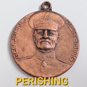 May include: A circular copper-toned metal medallion featuring a relief portrait of General John J. Pershing in a military uniform and cap. The text "GEN. JOHN J. PERSHING" is embossed along the curved edge, with a small loop at the top for hanging.