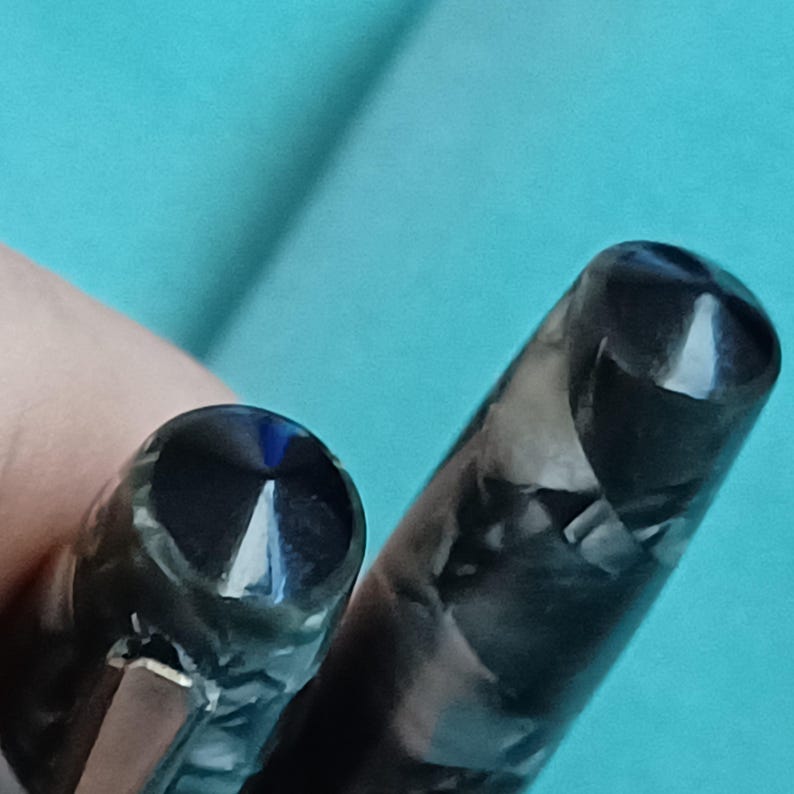 May include: Close-up of two fountain pens. The pen caps are black with a marbled pattern of gray and white. One pen cap has a blue accent. The pens are set against a light blue background.