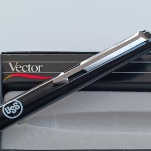 May include: A black pen with a silver clip and a silver band near the tip. The pen has a white logo that says "USS" on the side. The pen is in a black box with the word "Vector" printed on the top.