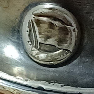 May include: Close-up of a silver coin with a raised image of a flag. The flag is a simple design with a single stripe.