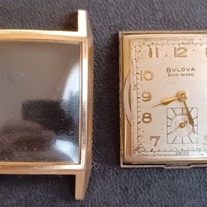 K&ouml;nnte beinhalten: Eine goldfarbene Armbanduhr mit quadratischem Zifferblatt und wei&szlig;em Zifferblatt. Das Zifferblatt der Uhr hat schwarze r&ouml;mische Ziffern als Stundenmarkierungen und schwarze Zeiger. Das Zifferblatt der Uhr hat auch den Text "Bulova Duo Wind" aufgedruckt. Die Uhr befindet sich in einem goldfarbenen Geh&auml;use mit einem klaren Glas.