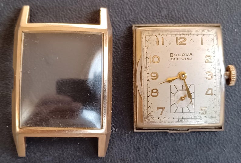 K&ouml;nnte beinhalten: Eine goldfarbene Armbanduhr mit quadratischem Zifferblatt und wei&szlig;em Zifferblatt. Das Zifferblatt der Uhr hat schwarze r&ouml;mische Ziffern als Stundenmarkierungen und schwarze Zeiger. Das Zifferblatt der Uhr hat auch den Text "Bulova Duo Wind" aufgedruckt. Die Uhr befindet sich in einem goldfarbenen Geh&auml;use mit einem klaren Glas.