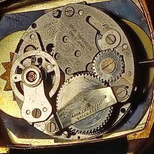 Puede incluir: Primer plano de un mecanismo de reloj con intrincados engranajes y componentes. Las piezas met&aacute;licas son plateadas y doradas, con texto visible que incluye "SWISS UNADJUSTED ONE 1 JEWEL" y "ADOR CHRONOMATIC WATCH CO". El reloj tiene una est&eacute;tica vintage.