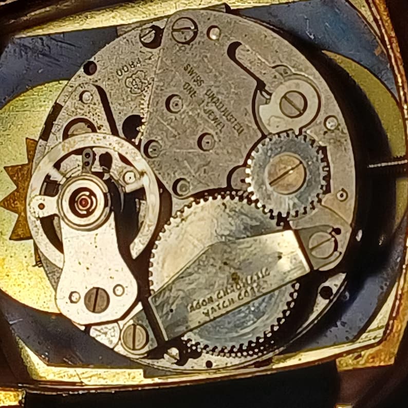 Puede incluir: Primer plano de un mecanismo de reloj con intrincados engranajes y componentes. Las piezas met&aacute;licas son plateadas y doradas, con texto visible que incluye "SWISS UNADJUSTED ONE 1 JEWEL" y "ADOR CHRONOMATIC WATCH CO". El reloj tiene una est&eacute;tica vintage.