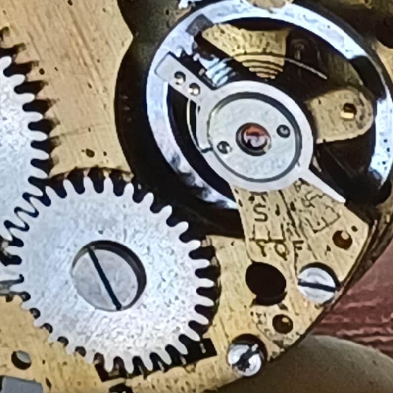 May include: Close-up of a vintage watch mechanism. The image shows intricate details of the internal gears, screws, and balance wheel. The gears are silver and gold, with a focus on the precision engineering of the timepiece.