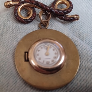 May include: A gold-toned brooch featuring a braided dark brown leather snake design at the top, with a circular gold-toned pendant hanging from it. The pendant contains a small analog watch face with a white background, gold hands, and gold numerals.