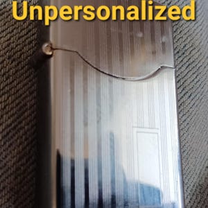 May include: A rectangular silver-tone metal lighter with a hinged lid and a vertical striped pattern. The word Unpersonalized is printed in yellow text across the top portion of the lighter. The surface is reflective with a smooth metallic finish.