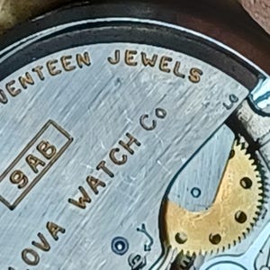 K&ouml;nnte beinhalten: Nahaufnahme eines silbernen Zifferblatts mit dem Text "Nova Watch Co." und "Seventeen Jewels" in goldener Schrift. Das Zifferblatt hat auch ein kleines Quadrat mit dem Text "9AB" darin.